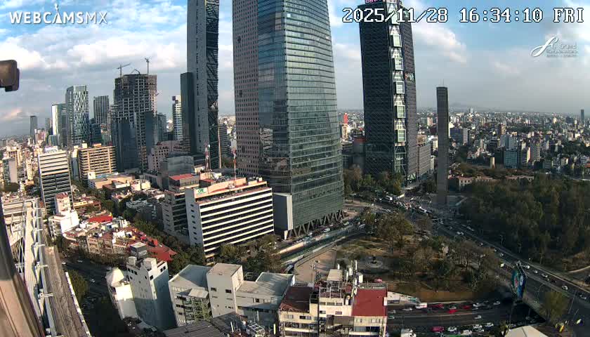 An expansive urban landscape unfolds from an elevated perspective, showcasing a mix of modern glass skyscrapers, bustling roads with traffic, and lower-rise buildings, all beneath a partly cloudy sky.