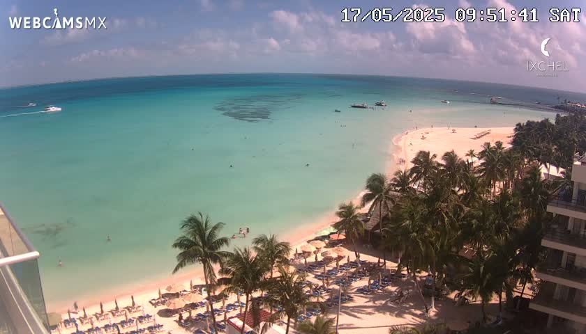 A beach with turquoise water, palm trees, beach chairs, and boats in the distance under partly cloudy skies.