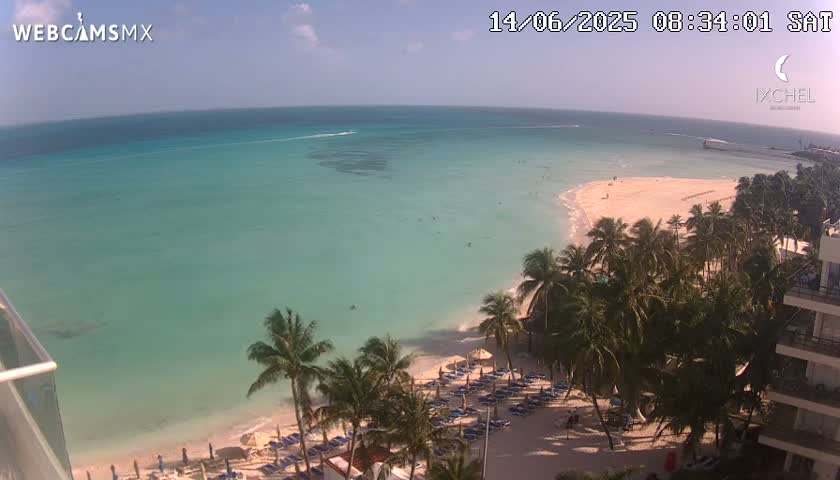 An aerial view shows a tropical beach with calm, clear turquoise water, a sandy shoreline lined with palm trees and beach umbrellas, and a section of a high-rise building in the lower right corner on a sunny day.
