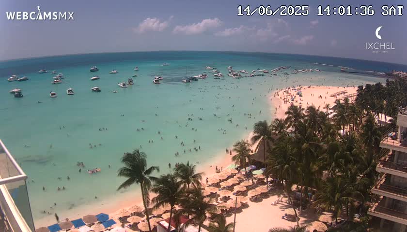 An aerial view shows a tropical beach with calm, clear turquoise water, a sandy shoreline lined with palm trees and beach umbrellas, and a section of a high-rise building in the lower right corner on a sunny day.
