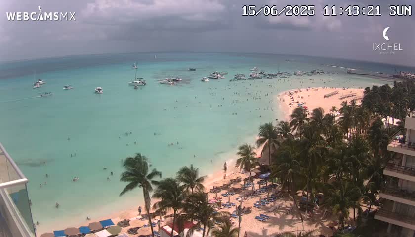 An aerial view shows a beach with many people swimming and sunbathing under partly cloudy skies, with numerous boats anchored in the calm, clear turquoise water.