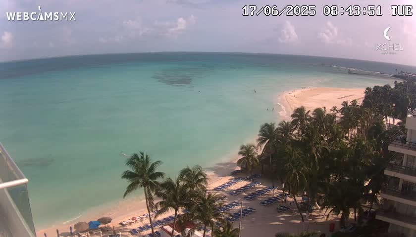 A partly cloudy day reveals a tranquil beach scene with turquoise water, white sand, palm trees, and lounge chairs lining the shore.