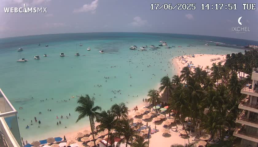 An aerial view shows a tropical beach with clear turquoise water, numerous people swimming and sunbathing, boats anchored offshore, and palm trees lining the shore under a partly cloudy sky.