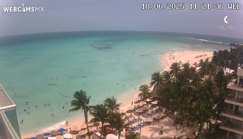 A beach with calm, turquoise water is seen from an elevated vantage point, with many people swimming and sunbathing, surrounded by palm trees and beach umbrellas under a mostly clear sky.