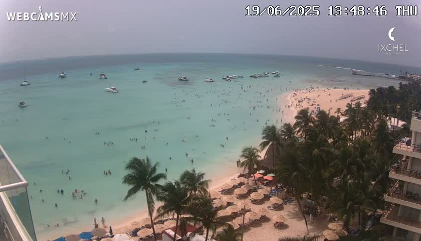 A beach with many people swimming and sunbathing under an overcast sky, numerous boats are visible in the calm, shallow turquoise water.