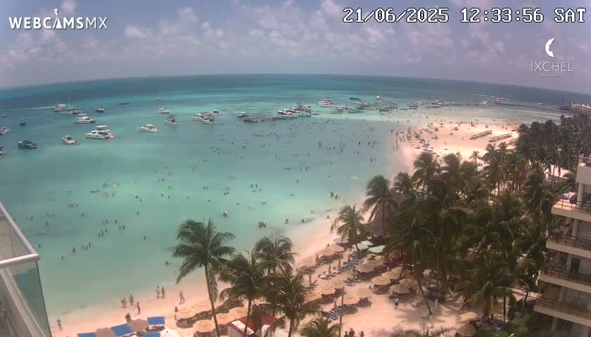 A partly sunny day shows a beach with many people swimming in clear, shallow turquoise water, numerous boats anchored offshore, and palm trees lining the shore.