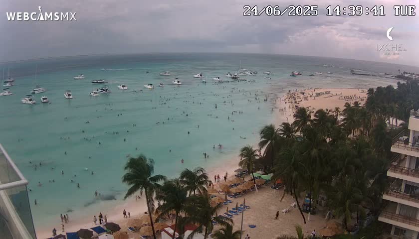 An aerial view shows a beach with many people swimming and sunbathing under an overcast sky, numerous boats are in the calm, turquoise water, and palm trees line the shore near a resort.