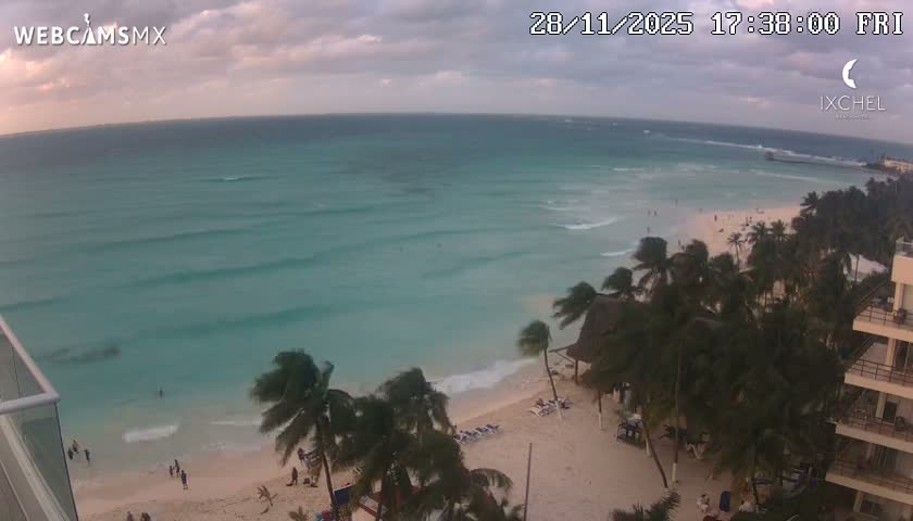 An aerial view captures a wide turquoise ocean with gentle waves meeting a sandy beach dotted with people, swaying palm trees, and resort buildings under a cloudy sky.