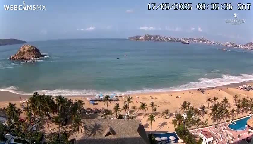 A sandy beach with palm trees and a resort is visible, with a calm ocean extending to a distant town under a partly sunny sky.