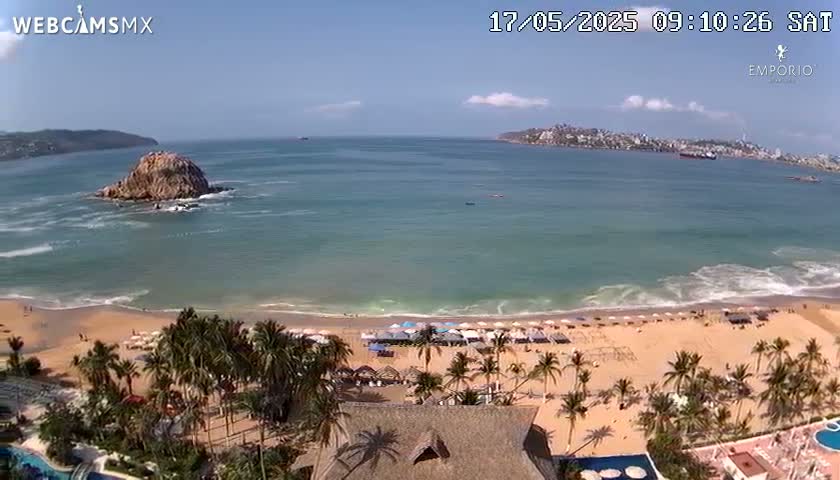 A sandy beach with palm trees and numerous umbrellas is visible beneath a partly cloudy sky, overlooking a calm ocean with a small island and a distant coastline.