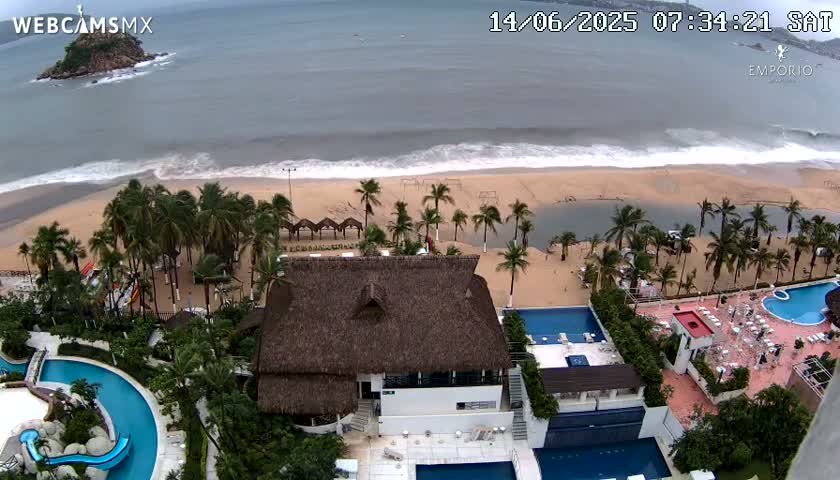 An aerial view shows a beach with moderate waves under an overcast sky, palm trees, resort buildings with multiple pools, and a small island in the distance.