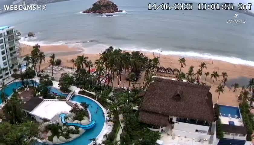 An aerial view shows a beach with moderate waves under an overcast sky, palm trees, resort buildings with multiple pools, and a small island in the distance.