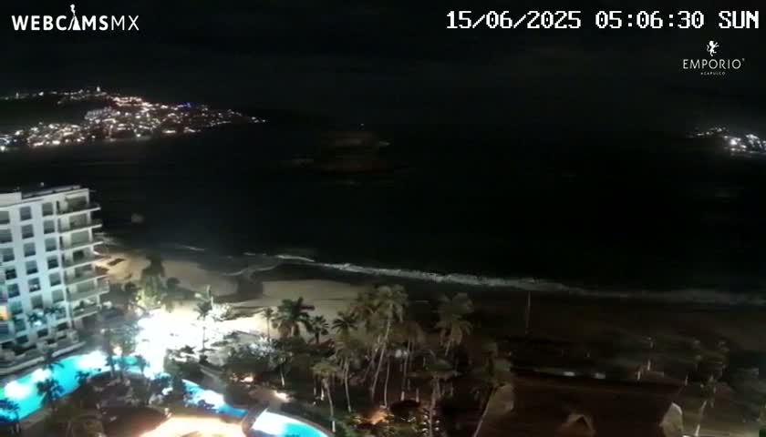 A nighttime view of a beach with gentle waves, a resort with pools, and a city illuminated on a distant hill.