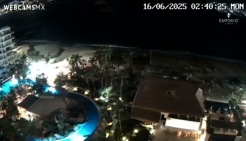 A nighttime aerial view shows a resort complex with illuminated swimming pools, palm trees, and a beach under a dark sky.
