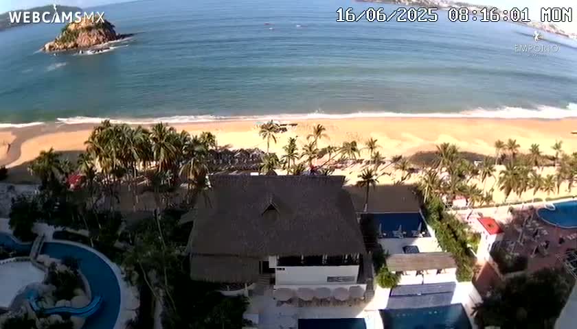 An aerial view shows a resort with palm trees, swimming pools, and a thatch-roofed building overlooking a sandy beach and calm ocean under a sunny sky.