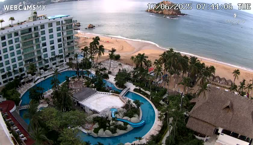 An aerial view shows a resort complex with multiple pools, a sandy beach, palm trees, and calm ocean water under a mostly clear sky.