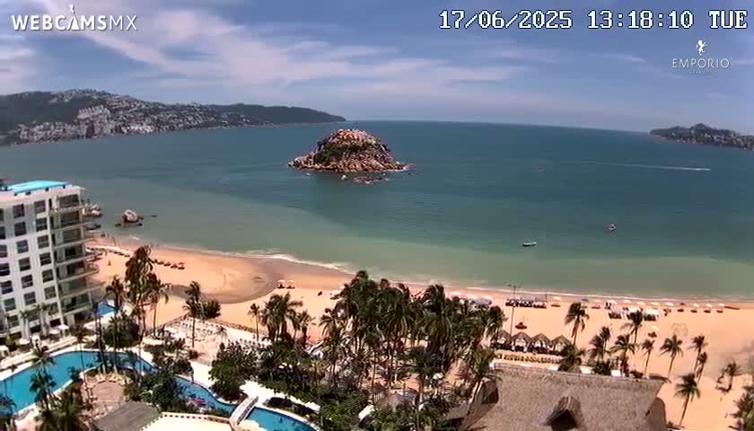 A sandy beach with palm trees and a resort complex borders a calm, clear ocean with a small island visible in the distance under a sunny sky.