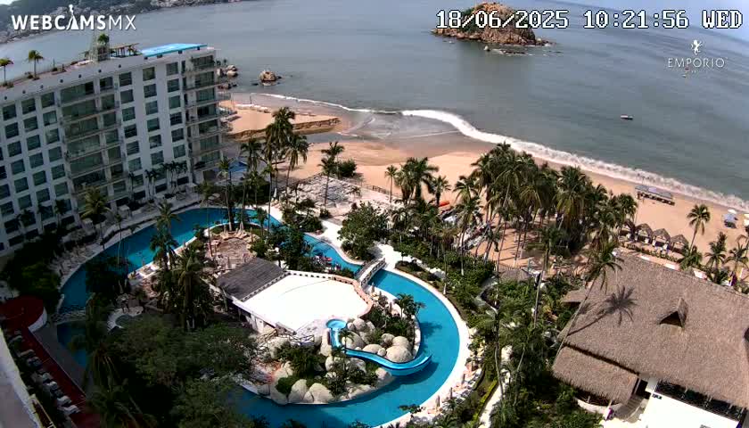 An aerial view shows a resort with a large, winding pool complex next to a sandy beach with palm trees and calm ocean waves under a sunny sky.