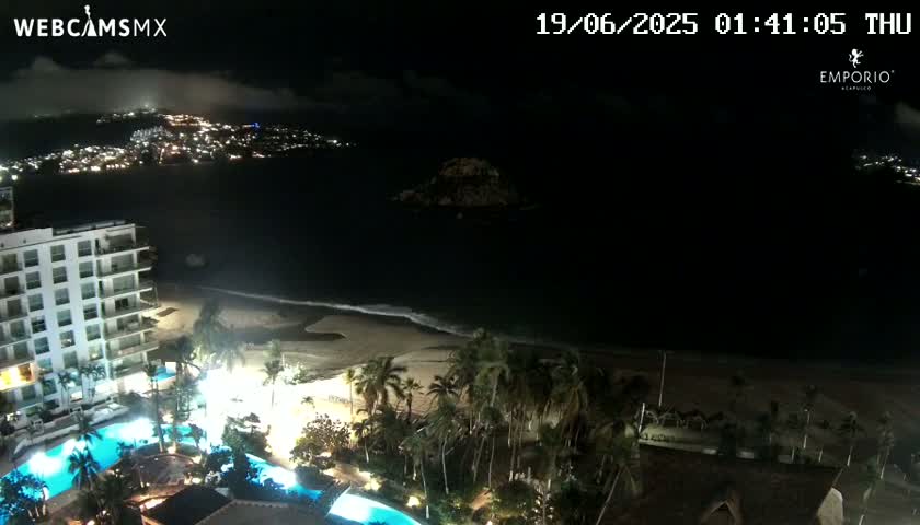 A nighttime view of a resort with pools and palm trees, situated on a beach near a city illuminated across a dark body of water under a partly cloudy sky.