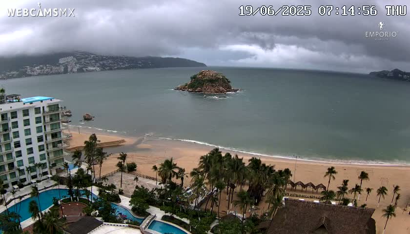 An overcast day shows a sandy beach with palm trees, a resort with pools, and a small island in a bay, with mountains in the background.