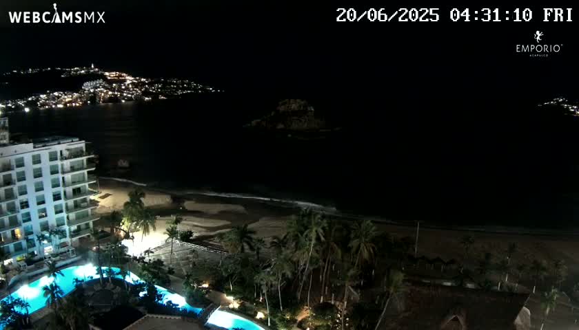 A nighttime view of a beach with a resort featuring swimming pools, palm trees, and a city skyline visible in the distance under clear skies.