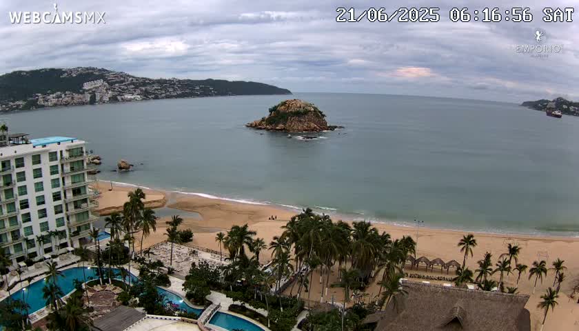 A mostly cloudy day reveals a sandy beach lined with palm trees, a resort with swimming pools, and a small island in a calm bay next to a mountainous coastline.