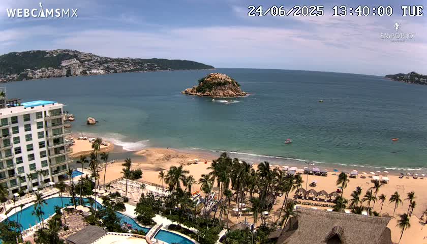 A beach resort with multiple pools overlooks a calm ocean and sandy beach dotted with palm trees, a small island, and distant mountains under a mostly sunny sky.