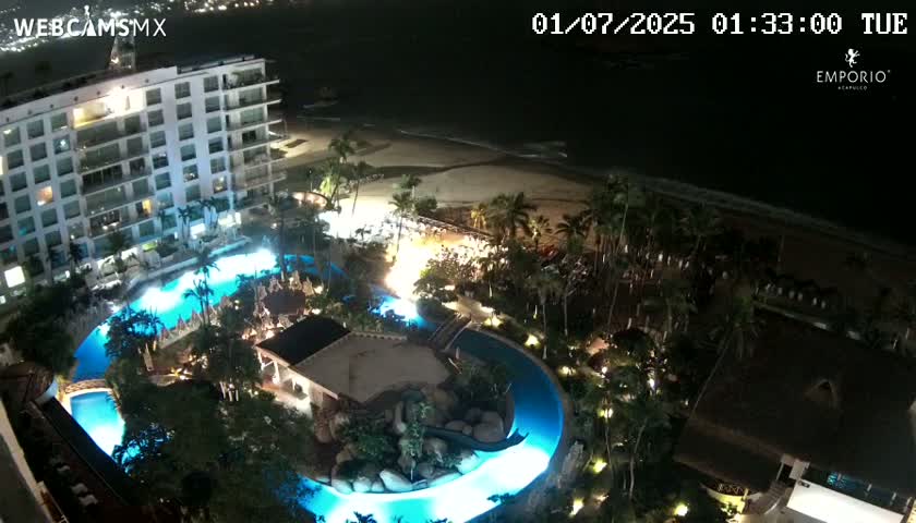 A nighttime aerial view shows a resort with a large, illuminated winding pool, surrounded by lush landscaping, a white building, and a dark beach and ocean beyond.