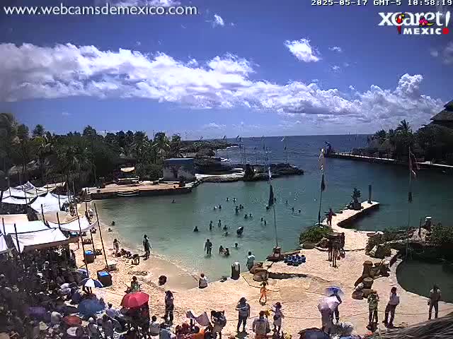 A partly sunny day at a tropical beach cove shows people swimming and relaxing on the sand near various structures and vegetation.