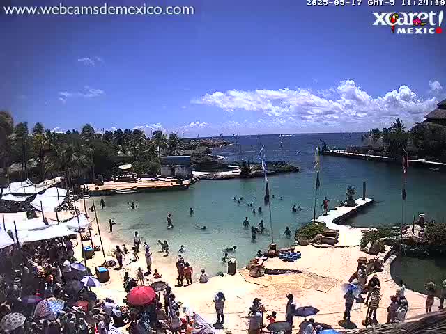 A sunny day at a tropical beachside resort with calm water, people swimming and lounging on the sand, and lush green vegetation surrounding a lagoon.