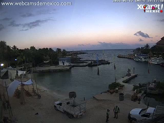 A tranquil waterfront scene at dusk shows a calm body of water with boats docked near a sandy shore, featuring some structures and vehicles on land.