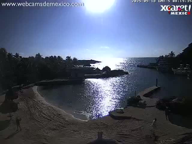 A sunny day reveals a secluded cove with calm water, a sandy beach, tropical vegetation, and a few people visible near the shoreline.