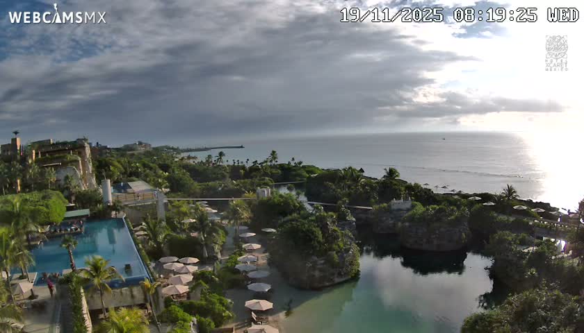 A partly cloudy sky overlooks a dark, calm body of water nestled between a sandy beach and silhouetted tropical vegetation.