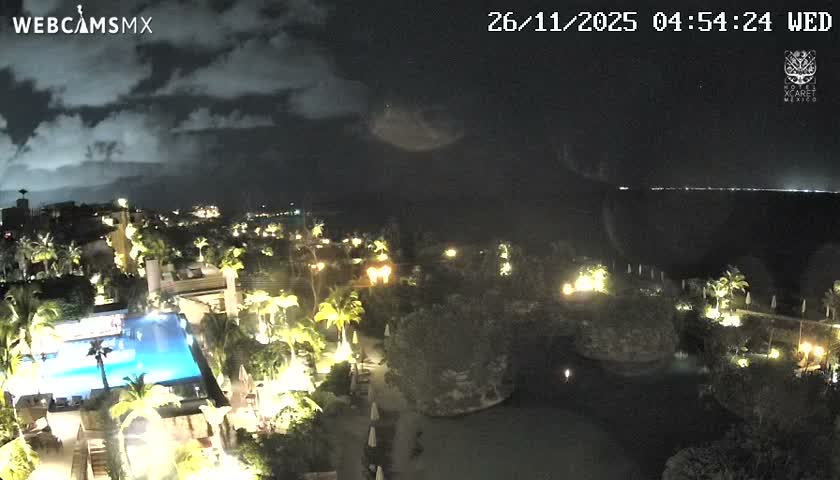 A nighttime view of a brightly lit resort complex reveals a sparkling blue swimming pool, numerous palm trees, illuminated buildings, and the dark ocean stretching into the distance under cloudy skies.