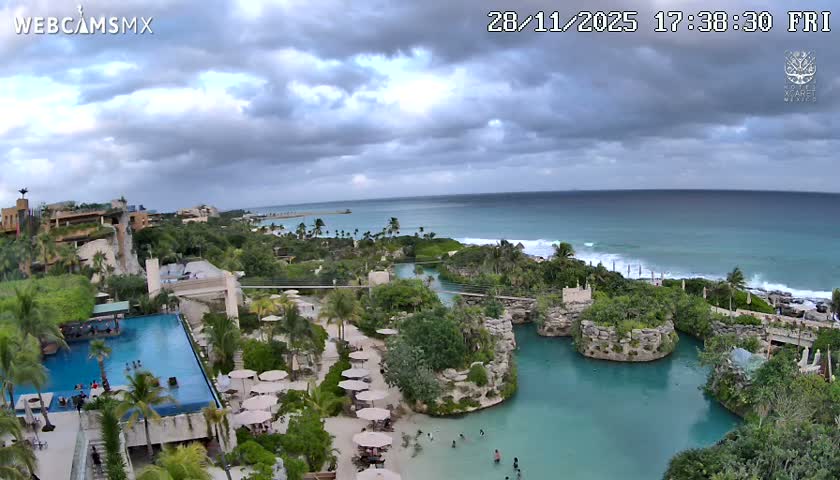 A wide aerial view captures a vibrant tropical resort area with large swimming pools, a natural turquoise lagoon with people swimming, and an ocean coastline featuring breaking waves under a heavily overcast sky.