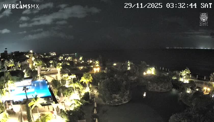 This night-time view captures a brightly lit resort complex with a swimming pool, lagoon, palm trees, and buildings, set against a cloudy sky and the dark ocean with distant lights on the horizon.