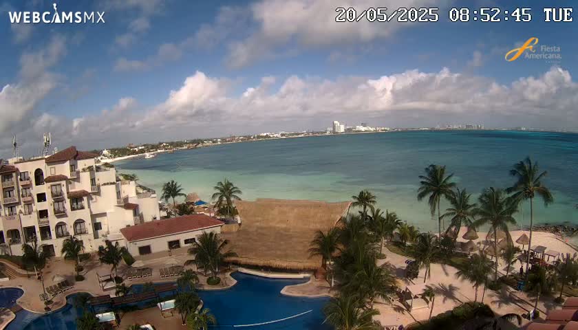 An aerial view shows a resort with swimming pools and palm trees bordering a calm, light-blue ocean under a partly cloudy sky.