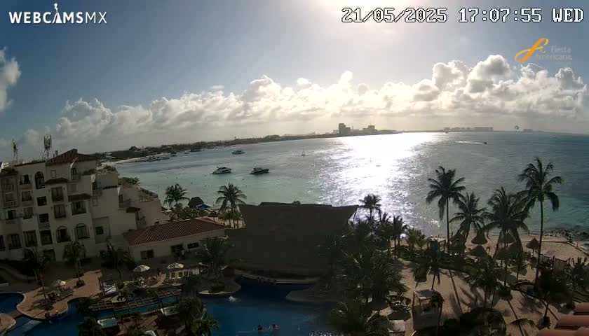 A partly cloudy afternoon overlooks a resort with pools and palm trees next to a calm ocean with several boats.