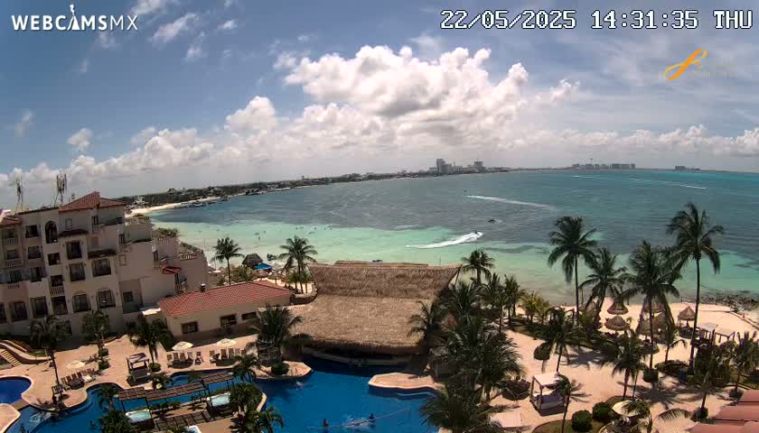 A resort with swimming pools overlooks a calm, light-blue ocean under a partly cloudy sky.