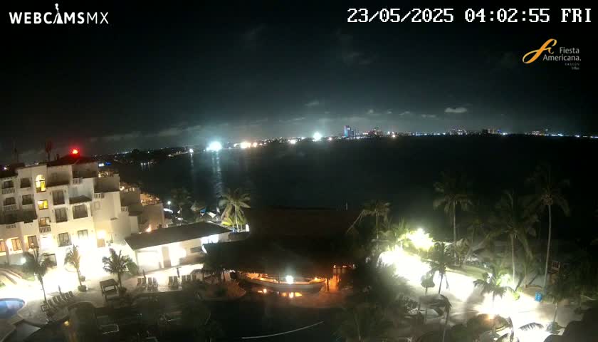 A nighttime, high-angle view shows a resort complex illuminated by artificial light, with a body of water and a distant city skyline visible under a partly cloudy sky.