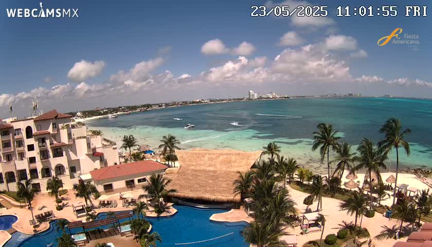 An aerial view of a resort with swimming pools, palm trees, and a beach overlooking a calm, turquoise ocean under a partly cloudy sky.