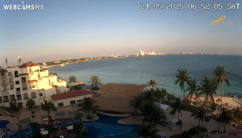 A resort overlooks a calm ocean at dawn, with palm trees lining the beach and a city visible in the distance.
