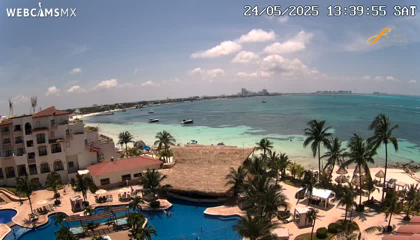 An aerial view of a resort on a sunny day, showing a beach, turquoise ocean, swimming pools, and palm trees under partly cloudy skies.
