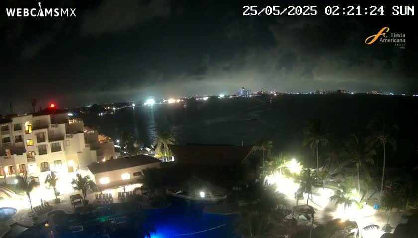 A nighttime view of a resort complex with pools, palm trees, and a city skyline visible across a dark body of water under a partly cloudy sky.