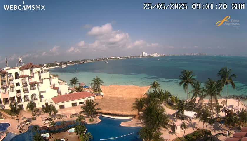 An aerial view of a resort on a sunny day shows a large beachfront hotel, swimming pools, palm trees, and a turquoise ocean with a city skyline in the distance.