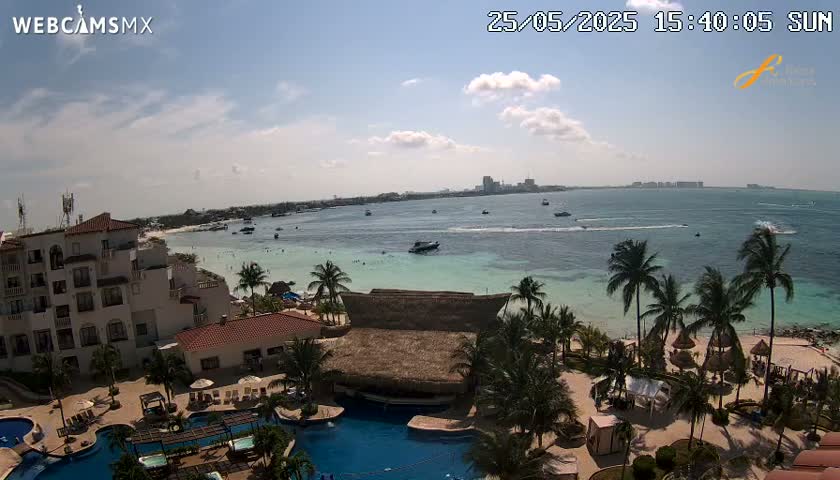 A sunny day at a beachfront resort shows a turquoise ocean, white sand beach, palm trees, resort buildings, swimming pools, and several boats in the distance.