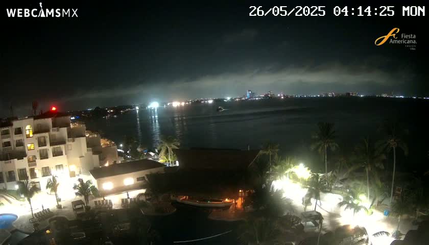 A nighttime view of a resort complex with pools and palm trees, overlooking a calm body of water with city lights visible in the distance under a cloudy sky.