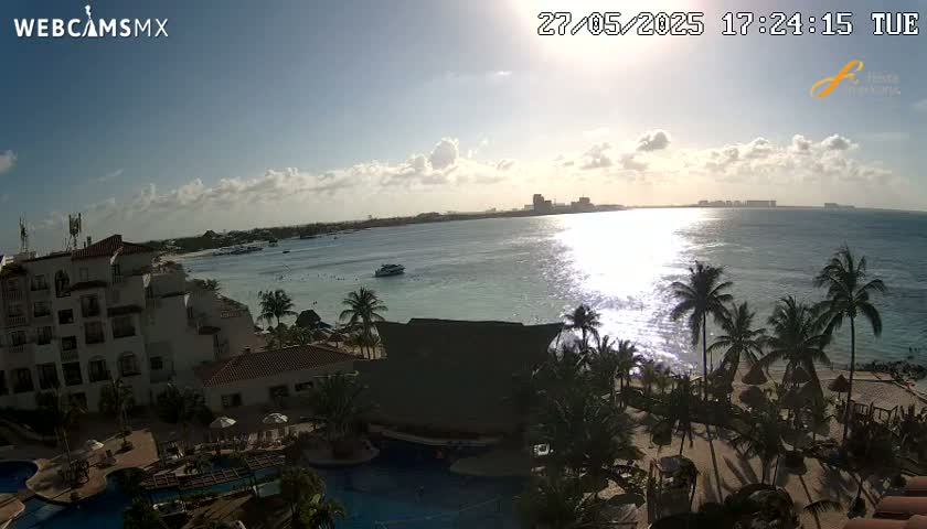 A partly sunny day overlooking a calm ocean with a resort and palm trees in the foreground.