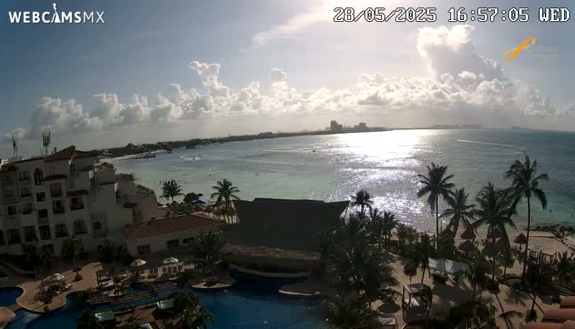 A partly sunny day shows a resort with swimming pools overlooking a calm ocean and a beach with palm trees.