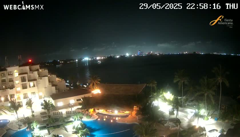A nighttime view of a resort complex with a swimming pool, illuminated buildings, and palm trees, overlooking a calm body of water and a distant city skyline under a clear sky.